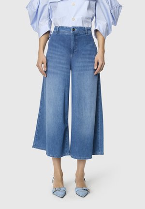 Femme portant un jean large cropped bleu clair, une chemise rayée bleu clair avec des manches bouffantes et des talons pointus bleus.