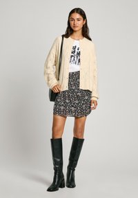 Cardigan a maglia crema, gonna nera a fiori, T-shirt bianca con stampa grafica, stivali neri sopra al ginocchio e piccola borsa a spalla nera.