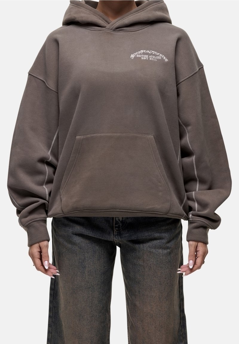 Ruskea ylikokoinen huppari, valmistettu pehmeästä kankaasta, jossa on etutasku, valkoinen logoteksti rinnassa ja kontrastiset ompelusaumat.