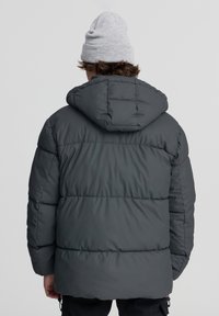 Szary puchowy płaszcz z poziomymi przeszyciami, kapturem i gładką, błyszczącą fakturą. Noszony z czarnymi spodniami i szarym beaniem.