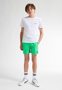 Bílý bavlněný tričko s malým logem, sp�árované s živě zelenými šortkami. Oblečení doplňují bílé tenisky a pruhované ponožky.