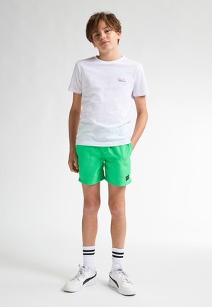 Camiseta de algodón blanca con un pequeño logo, combinada con pantalones cortos verdes vibrantes. El atuendo se completa con zapatillas blancas y calcetines a rayas.