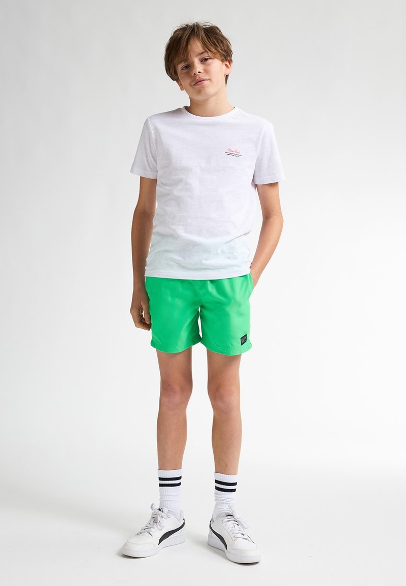 Bílý bavlněný tričko s malým logem, spárované s živě zelenými šortkami. Oblečení doplňují bílé tenisky a pruhované ponožky.