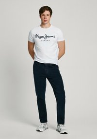 Bijela majica kratkih rukava s natpisom "Pepe Jeans London" ispisanim tamnoplavim slovima, uparena s tamnoplavim hlačama i sivim sportskim cipelama.