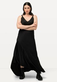 Robe maxi noire en satin avec un décolleté en V, des bretelles ajustables et un ourlet asymétrique. Associée à des bottes noires à semelle épaisse. Texture lisse.