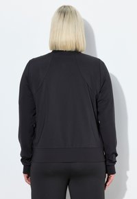 Giacca nera con zip, maniche lunghe, polsini e vita a coste. Tessuto liscio con un design aderente, mostrata da dietro.