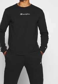 Langärmliges schwarzes T-Shirt mit Rundhalsausschnitt, das das weiße "Champion"-Logo auf der Brust und kontrastierende Streifen entlang der Ärmel aufweist.