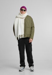 Olivgrüne gesteppte Jacke, weißer Fransen-Schal, schwarze Cargo-Hosen, lila Beanie und Sonnenbrille, kombiniert mit mehrfarbigen Sneakers.