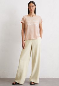 Lichtroze katoenen t-shirt met "Liefde, Vrede & Harmonie" in het wit gedrukt, gecombineerd met wijduitlopende lichtgele broek en zwarte sandalen.