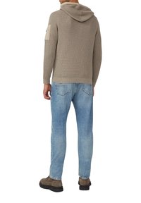 Beige Strick-Hoodie mit Kapuze, ausgestattet mit einer aufgesetzten Tasche am linken Arm. Kombiniert mit hellblauen Jeans und braunen Schuhen.