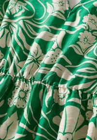 Tissu vert avec motifs floraux et feuillages blancs, avec une couture élastique froncée au centre pour un effet plissé.