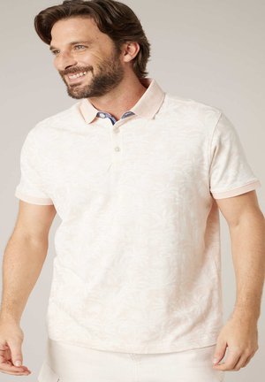 Man lacht terwijl hij een lichtroze poloshirt met patroon en een contrasterende binnenkraag draagt, staand tegen een effen achtergrond.