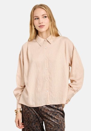 Joven mujer con una camisa beige suelta abotonada y pantalones oscuros con estampado, de pie con una mano en el bolsillo.