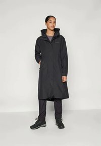 Črn vodoodporen plašč z nastavljivo kapuco, dolgimi rokavi, žepi z zadrgo in zategnjenim pasom. Nosa se nad temnimi hlačami in trdih čevljih.