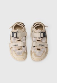 Zapatos de exterior beige con parte superior de malla, correas ajustables y suela de goma. Presentan una marca en las correas y un diseño texturizado único.