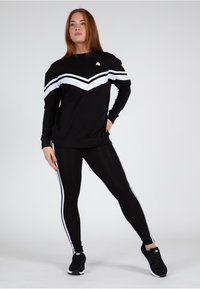 Felpa nera con strisce bianche a chevron, abbinata a leggings neri con strisce laterali bianche e scarpe da ginnastica nere.