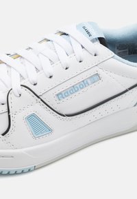 Fehér bőr sneaker kék részletekkel, textúrált oldalsó panellel, fekete díszítéssel és sík fehér fűzőkkel. Figyelemre méltó feltűntetett logó az oldalon.