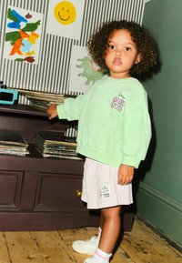 Menta zöld bő szabású pulóver "Daisy Duck" grafikával, világos rózsaszín csíkos szoknyával párosítva. Fehér tornacipők és színes zokni kiegészítők.