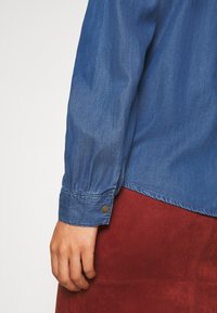 Personne portant une chemise bleue à manches longues boutonnée et une jupe rouge en daim, la main droite détendue le long du corps.