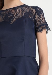 Marine jurk met een aansluitende lijfje en een kanten overlay op korte mouwen, met een golvende halslijn en textuurstof.