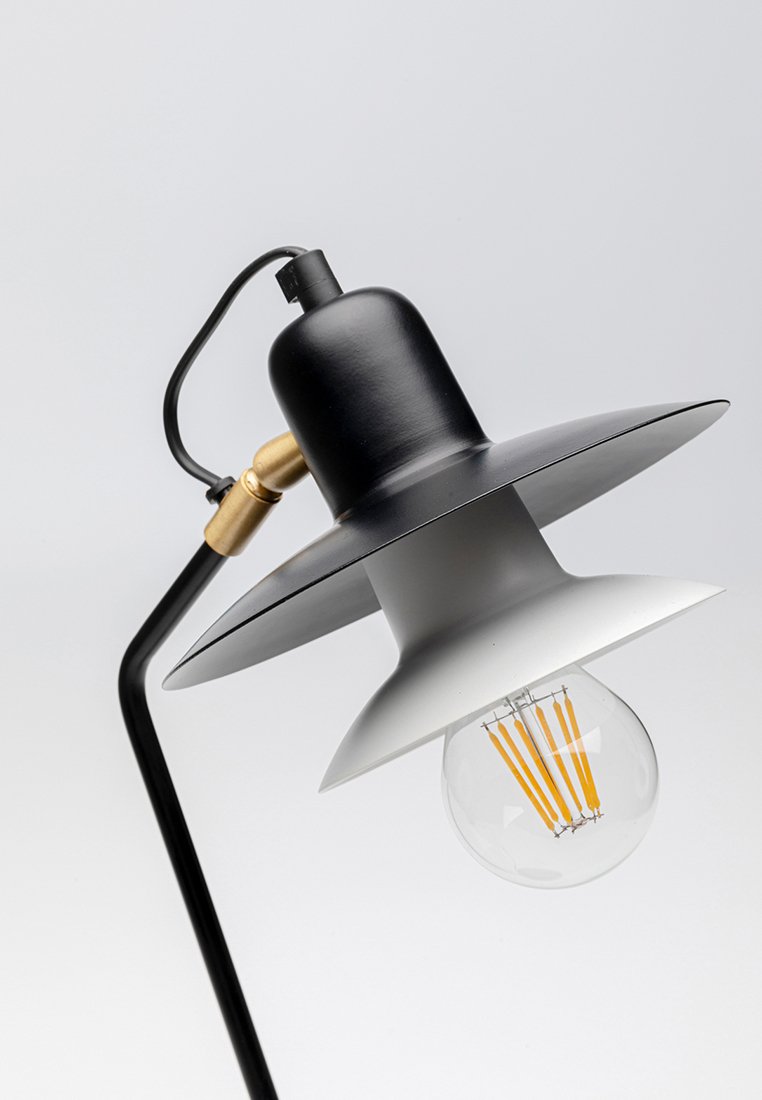 Lámpara de mesa con pantalla de metal negro, acento blanco y hardware de latón. Presenta una bombilla clara con filamento visible, sostenida por un tallo curvado negro.