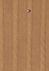 Maglione a maglia cable in un caldo colore beige, con un motivo testurizzato di cavi intrecciati e una piccola toppa con logo in alto a sinistra.