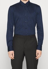 Granatowa koszula z geometrycznym niebieskim wzorem, zapinana na guziki, z kołnierzykiem; zestawiona z czarnymi spodniami, podkreślając styl smart casual.