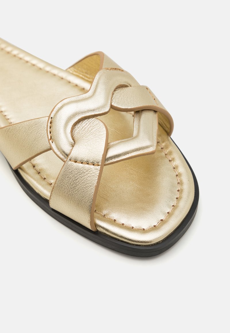 Goldene Leder-Sandale mit einem herzförmigen gewobenen Riemen-Design, genähten Details und einer flachen schwarzen Sohle. Glatte Textur mit metallischem Glanz.