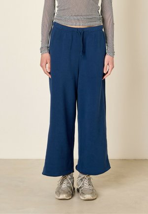 Person iført marineblå bukser med vidde ben og snøre i taljen, stribet langærmet crop top og sølvfarvede sneakers på en lys gulv.