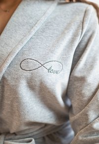 Accappatoio in morbido tessuto grigio, caratterizzato da un simbolo dell'infinito ricamato con la parola "amore" in corsivo. Include una chiusura con cintura e maniche ampie.