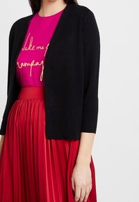 Kobieta nosząca czarny kardigan na magentowej bluzce z złotym napisem oraz spódnicy w kolorze czerwonym z wysokim stanem i plisami, stojąca na jednolitym tle.