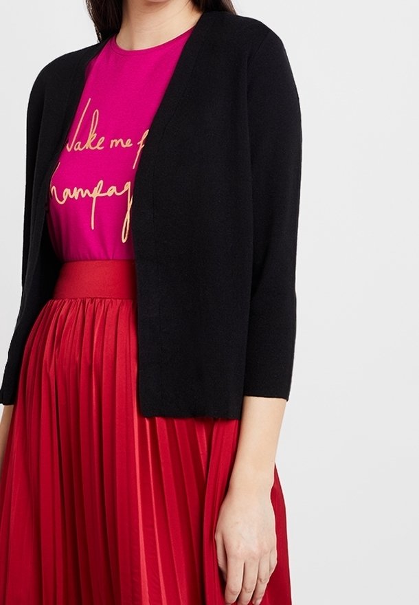 Kobieta nosząca czarny kardigan na magentowej bluzce z złotym napisem oraz spódnicy w kolorze czerwonym z wysokim stanem i plisami, stojąca na jednolitym tle.