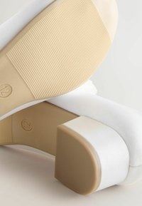 White heeled shoes with a textured beige sole, featuring a square heel. The material appears smooth with a matte finish and soft edge detailing.