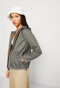 Gray lightweight jacket with a zip closure, featuring yellow and black stripes, hood, and side pockets, paired with a white bucket hat.
