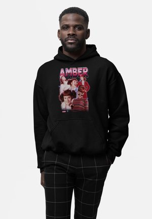 Mand iført sort hættetrøje med farverigt collage af en kvindes forskellige ansigtsudtryk og ordet "AMBER" med store bogstaver.