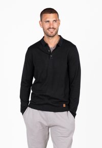 Polo a maniche lunghe nera con patta a tre bottoni, polsini a costine e tessuto testurizzato, abbinata a pantaloni jogging grigio chiaro. Etichetta in pelle sul fondo.