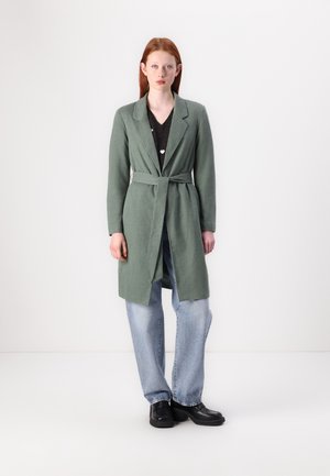 Light green long coat with a belted waist, notched lapels, and side slit pockets, worn over a dark top and loose denim jeans.