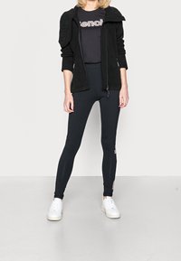 Chaqueta de forro polar negra con cremallera sobre una camiseta negra con el logo "Bench", combinada con leggings negros y zapatillas blancas, presentando un diseño minimalista.