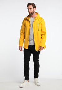 Chaqueta impermeable amarilla con capucha, con cierre frontal, bolsillos laterales y sudadera gris debajo. Combinada con pantalones negros y zapatillas blancas.