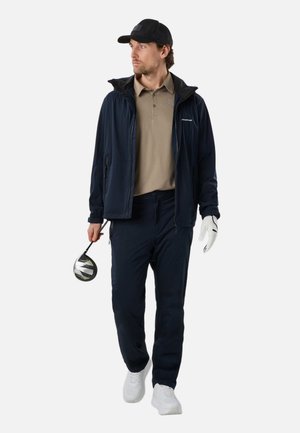 Man met een zwarte pet, marineblauwe jas, beige polo en marineblauwe broek, lopend met een golfclub in de rechterhand en een witte handschoen aan de linkerhand.