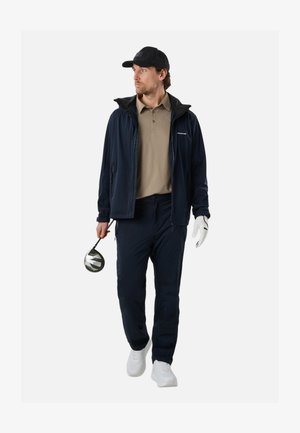 Man met een zwarte pet, marineblauwe jas, beige polo en marineblauwe broek, lopend met een golfclub in de rechterhand en een witte handschoen aan de linkerhand.