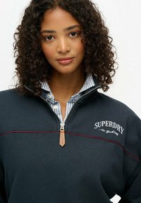 Sweat-shirt zippé marine avec des coutures rouges ; il a un col haut, un tirant en cuir pour la fermeture éclair et un logo brodé sur la poitrine.