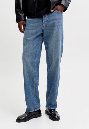 Personne portant un jean bleu clair ample, des chaussures en cuir noires et une veste en cuir noire, une main dans la poche.