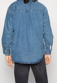 Uomo che indossa una camicia di jeans blu a maniche lunghe e jeans neri, mostrato di spalle su uno sfondo chiaro e semplice.
