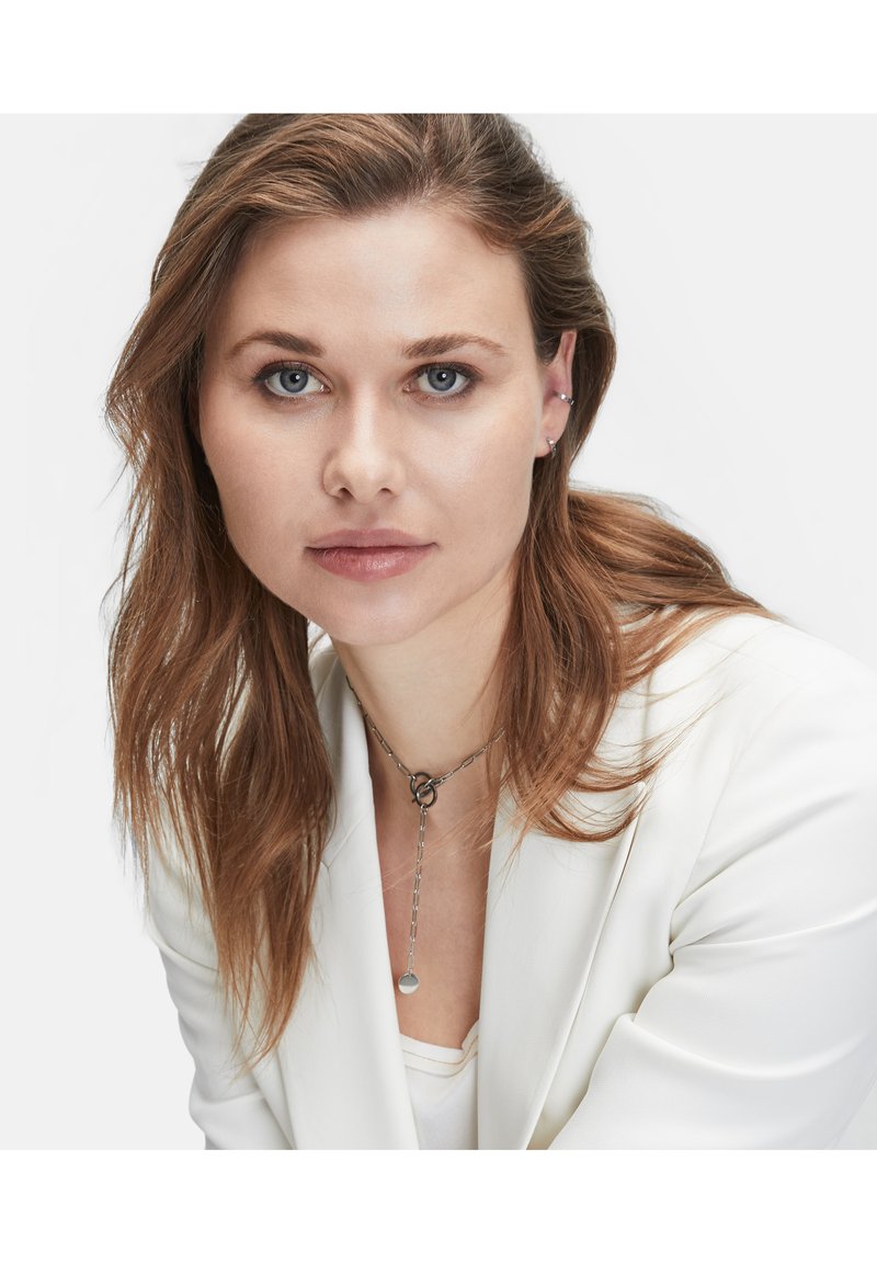 Silberne Halskette mit einem Kettendesign, das einen runden Anhänger zeigt. Modell trägt ein weißes Blazer und ein weißes Oberteil, mit schulterlangem braunem Haar.