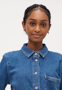 Denimska srajca v modri barvi z ovratnikom, gumbom spredaj in žepom na prsih. Šivanje subtilno kontrastira s teksturo tkanine.