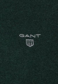 Tissu tricoté vert foncé avec un logo brodé blanc incluant le mot "GANT" et un design de bouclier avec trois étoiles.