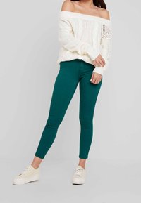 Leggings verdes elásticos combinados con un suéter de punto color crema, con hombros al aire, y zapatillas blancas. La ropa presenta un corte holgado y una telas texturizadas.