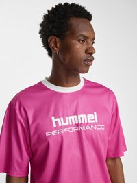 Jonge man met kort krullend haar die een roze "hummel PERFORMANCE" T-shirt met witte kraag draagt, kijkend naar rechts tegen een effen achtergrond.