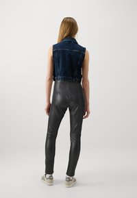 Jeansväst med ärmlös design, mörkblå färg. Tätt sittande svarta fuskläderbyxor med slät textur, i kombination med neutrala sneakers.